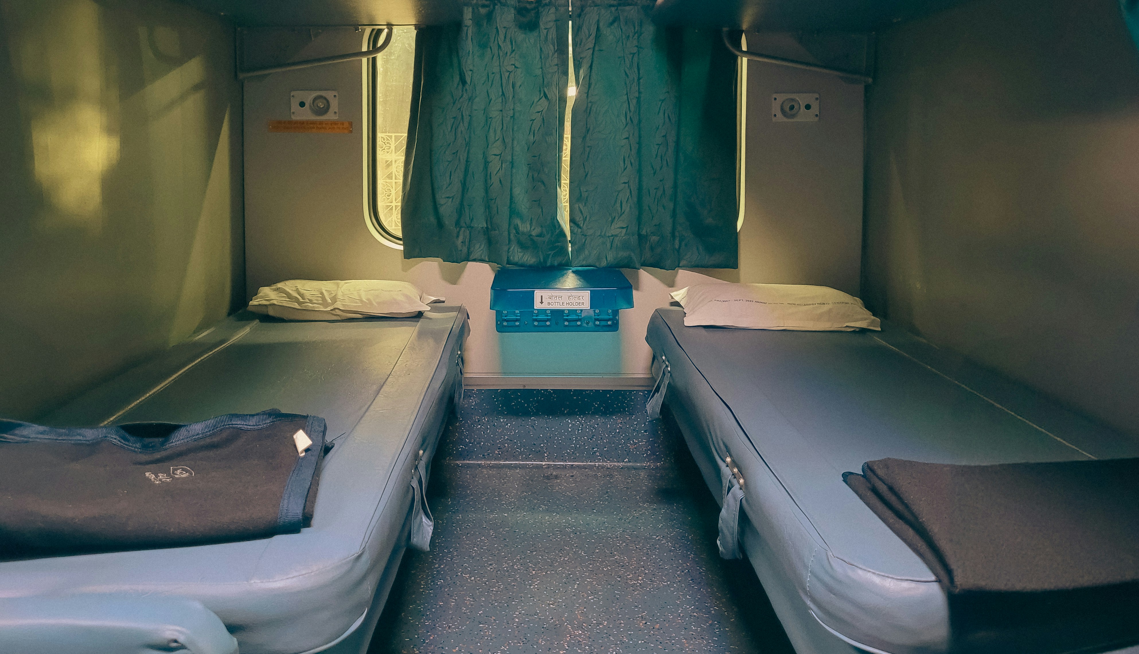 Inside view of a Sleeper Train in India showing two lower berths, pillows, blankets, and a window with curtains.